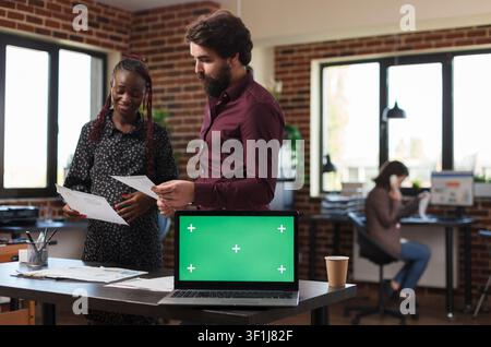 Marketing company office desk with green screen background laptop display on top of it. Pregnant woman and man looking at business paperwork having isolated background and mockup template on computer. Stock Photo