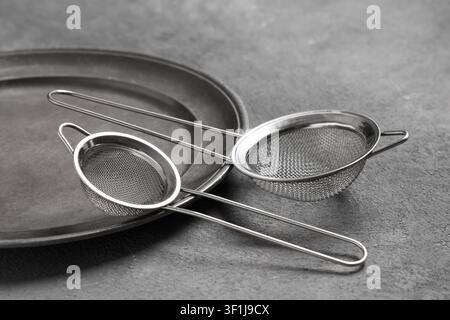 Two stainless steel mesh strainers and a metal plate on a dark gray concrete background, kitchen utensils and cooking tools Stock Photo