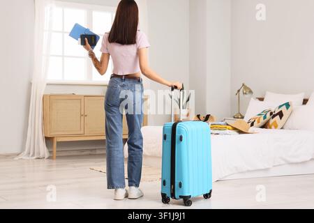 Young woman holding a passport and pulling a blue suitcase in a bright bedroom, ready for travel and vacation, Anticipation of a vacation or holiday Stock Photo