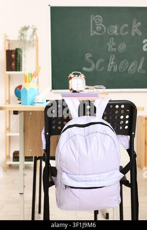Purple backpack hanging on a chair in a classroom with a blackboard displaying "back to school" text Stock Photo