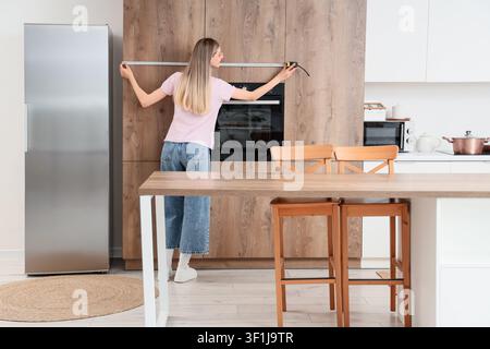 Young woman measuring kitchen cabinets with a tape measure for home renovation and interior design planning, the modern kitchen features wooden cabine Stock Photo