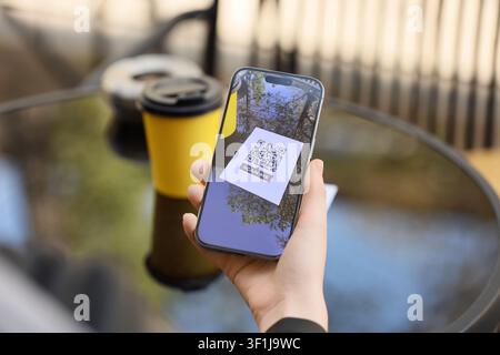 Hand holding a smartphone displaying a QR code with "scan me" text, digital payment and contactless technology concept, a yellow coffee cup is visible Stock Photo