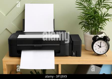Black inkjet printer with blank paper on a wooden shelf, home office and productivity concept, green wall background Stock Photo