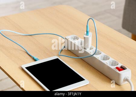 Tablet charging from a white power strip with a blue cable on a light wooden table, modern technology and energy, this setup modern technology energy Stock Photo