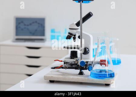 Laboratory setup with blue liquid in an erlenmeyer flask, microscope, and laptop on a white desk Stock Photo