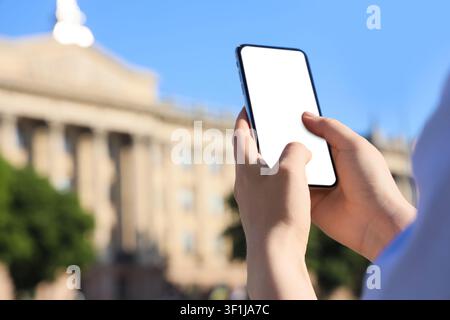 Person holding a smartphone with a blank white screen, interacting with a mobile device outdoors against a blurred building Stock Photo