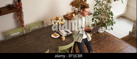 Handsome manager, man in glasses sits on table, holds laptop and types, sends message, works on project, drinks coffee, spends time at co-working Stock Photo
