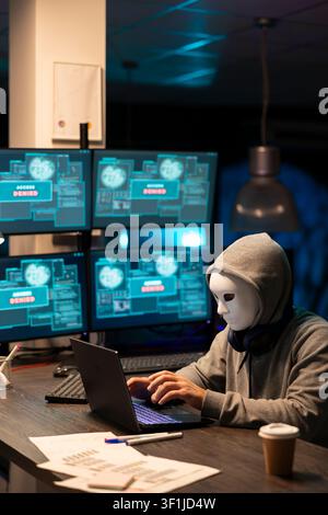 Hooded cybercriminal with concealed identity works late at night, typing malicious code on laptop. Masked man focuses on breaching security networks and accessing encrypted data in dark office. Stock Photo