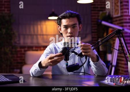 Male influencer holding headphones while speaking to camera and podcast microphone in home studio. Asian man produces lifestyle and technology content for social platforms and live streaming episodes. Stock Photo