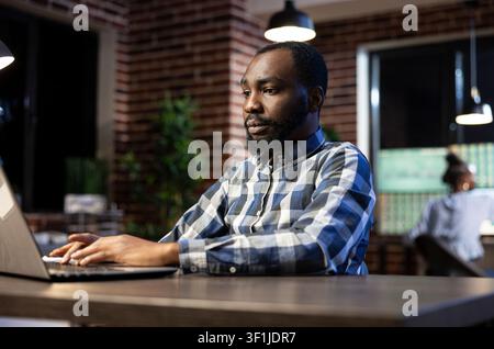 Financial analyst studying stock data on laptop capturing male financial analyst works late at a brick wall office desk, reviewing documents and stock data on his laptop. Stock Photo