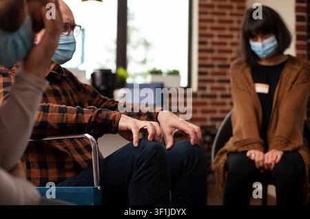 People wearing face masks sit in a circle before an addiction recovery discussion, preparing for counseling and therapist guidance in a group therapy room. Stock Photo