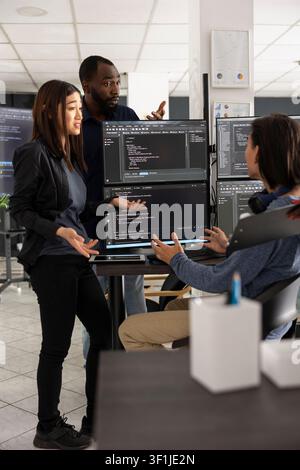 Diverse developers discuss programming data and source code on desktop pc during team meeting. Colleagues collaborate closely, brainstorming system improvements and digital problem solving strategies. Stock Photo