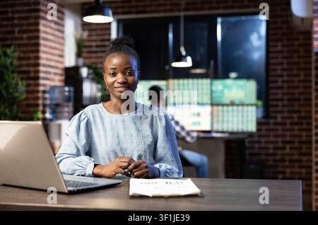 Black female stock analyst works late with a laptop and clipboard at her desk, looking confidently toward the camera in a trading office. Stock Photo