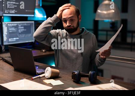 Bearded it specialist holds sheets of paper beside laptop as digital monitors show active code. The scene conveys technical workload, problem solving and cybersecurity documentation review. Stock Photo