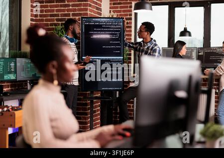 African American system engineers analyze source code for errors while checking a large monitor in an IT agency office focused on cybersecurity. Stock Photo
