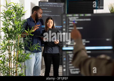 African american and asian programmers collaborate on laptop, comparing code to improve company website. Diverse colleagues work together in modern office, reviewing HTML scripts for web development. Stock Photo