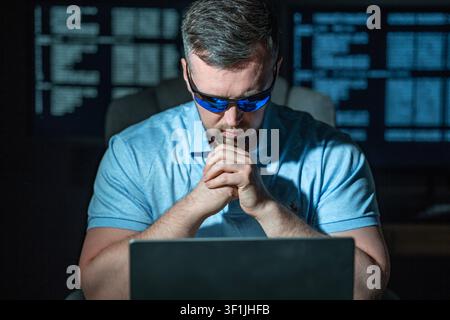 Hacker operates computer during night work. Hacker analyzes system data and logs. Hacker monitors cyber threats in network. Hacker performs digital Stock Photo