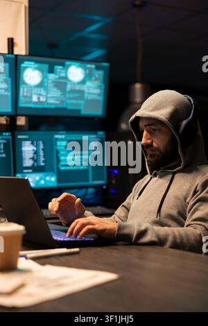 Hooded programmer monitors online activity, preventing hacking attempts and maintaining information security. White man works late in tech office, writing coding scripts for catching fraud and scammers Stock Photo