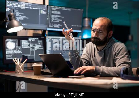 Developer stares at laptop with confused expression while holding pen and working on programming data. Multiple monitors display complex code, emphasizing challenges of technical decision making. Stock Photo