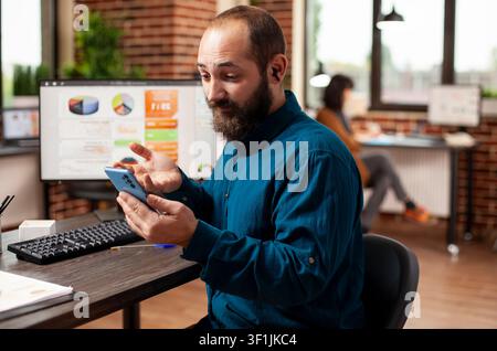 Bearded employee uses a smartphone for an online video call at an office desk, connecting mobile communication with everyday business work. Stock Photo