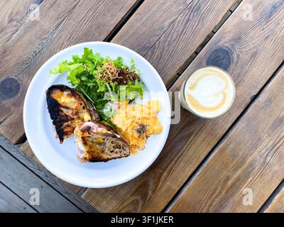 A white plate with toasted sourdough bread, scrambled eggs, and a mixed green salad is accompanied by a glass of latte with latte art, photographed from overhead on a wooden table. Stock Photo