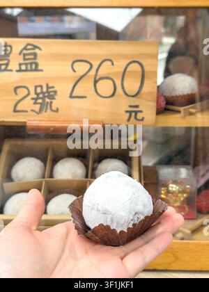 A hand holds a round white strawberry daifuku mochi on a brown paper cupcake liner in front of a bakery display case at Dihua Street in Taipei, Taiwan, with a price sign visible in the background. Stock Photo