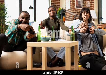 African american and caucasian couples play video games at home pointing at device screen, sharing snacks and drinks. Diverse friends laugh, smile and bond during online gaming in cozy apartment. Stock Photo