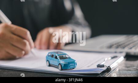 Buyer signs car loan purchase finance vehicle contract document, securing insurance and ownership with small blue toy model on clipboard while working Stock Photo