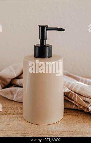 A matte ceramic soap dispenser with a black pump sits on a wooden countertop next to folded striped linen towels in a warm, minimalist neutral-toned s Stock Photo