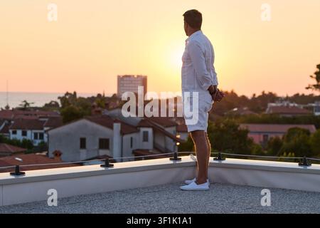 Man Standing on Rooftop Terrace Watching Sunset Over Seaside Town From Home Balcony Stock Photo