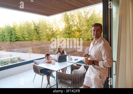 Family On Modern Balcony Terrace Having Coffee And Using Laptop While Father Stands At Door Stock Photo