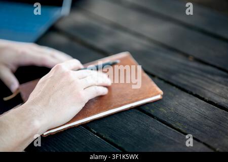 Hands resting on brown notebook with pen on dark wooden table, outdoor workspace concept. Stock Photo