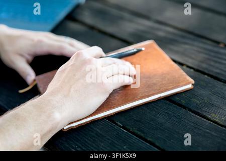 Hands resting on brown notebook with pen on dark wooden table, outdoor workspace concept. High quality photo Stock Photo