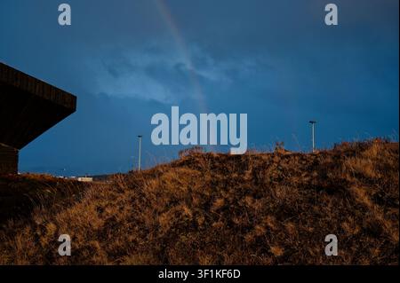 A faint rainbow arcs across a moody, dark sky over golden grassy hills Stock Photo