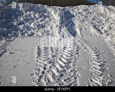 Tracks from tires cut through the snow with large snow piles in the background under clear sky during winter. Stock Photo