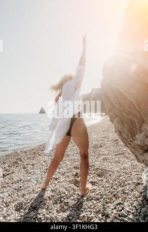 Woman beach vacation enjoying a sunny day on a pebbled shore while wearing a white shirt and black bikini Stock Photo