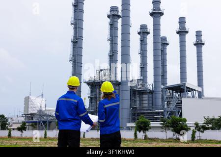Two professional engineers in safety gear examine a site plan document outside an industrial power plant for maintenance and construction project plan Stock Photo