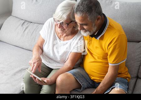 Happy senior couple sitting together on the sofa while looking at a smartphone. Smiling as they browse online, communicate, or share digital content, Stock Photo