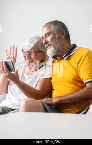 Happy senior couple sitting together on the sofa while making a video call on a smartphone. Stock Photo