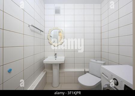 bright bathroom features white tile walls, sink, toilet, and washing machine. A decorative mirror hangs above the sink. Ventilation grill on the wall Stock Photo