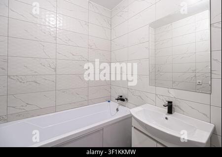 modern bathroom with white marble-like tiles, a white bathtub, a sleek sink with a black faucet, and a large mirror Stock Photo