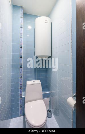 small bathroom with light blue tiles, a white toilet, and a water heater mounted on the wall Stock Photo