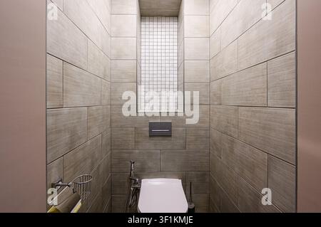 modern, tiled bathroom. The walls are covered in light brown, wood-look tiles, with a rectangular niche filled with white mosaic tiles. A toilet is ce Stock Photo