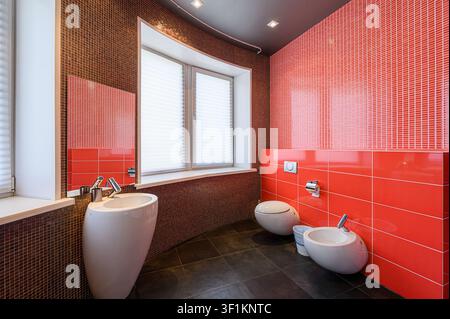 Modern bathroom featuring red & brown tiled walls, black floor, and sleek fixtures. Window with white blinds provides light Stock Photo