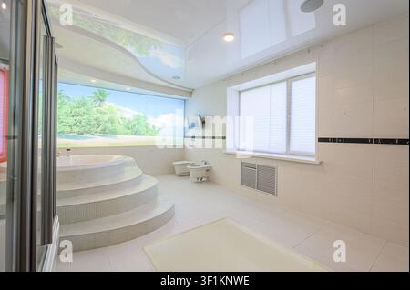 Bright bathroom with a jacuzzi tub, tropical mural, and a window with blinds. All in white and cream Stock Photo