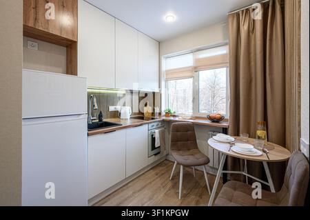 bright kitchen featuring white cabinets, wooden countertops, and a small dining area with brown chairs. Sunlight streams through the window, illuminat Stock Photo