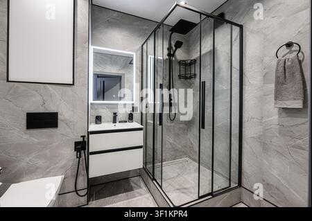 Modern bathroom with grey marble tiles, a black-framed glass shower, a white vanity with black fixtures, and a lighted mirror. Clean and minimalist de Stock Photo