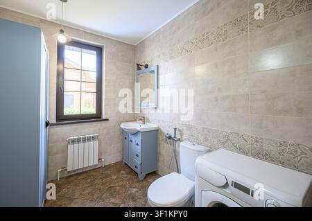 well-lit bathroom featuring beige tile walls, a blue vanity, a white toilet, and a washing machine. A window adds natural light Stock Photo