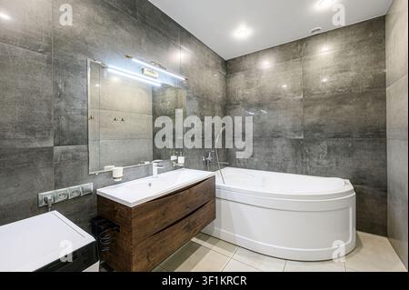 modern bathroom featuring gray tile walls, a white bathtub, and a dark wood vanity with a white sink. A mirror and lighting fixture are above the sink Stock Photo