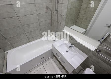 modern bathroom features gray tiled walls, a white bathtub with shower, and a sleek white vanity with a large mirror above. The flooring is also light Stock Photo